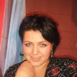 Uśmiechnięta kobieta z krótkimi, ciemnymi włosami i kolczykami, pozująca na tle łazienki z czerwonymi płytkami i zasłoną prysznicową.